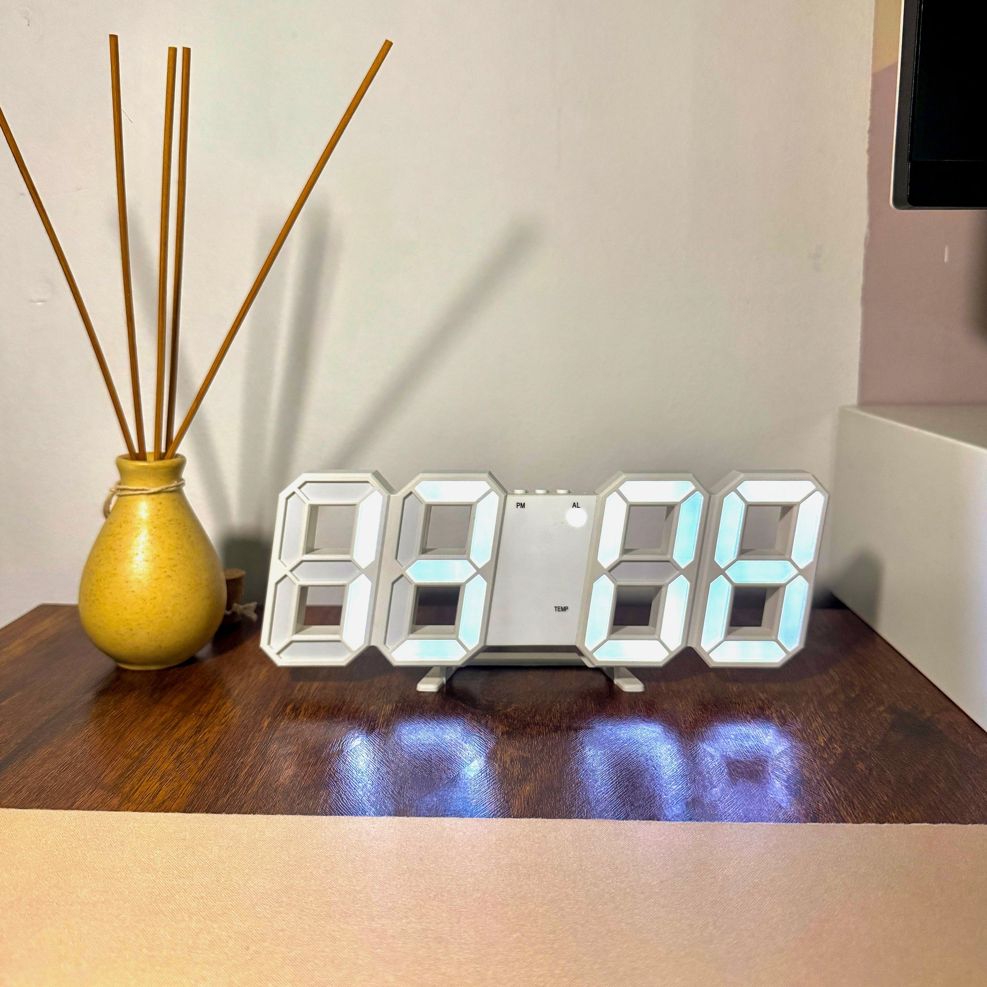 Digital clock on a wooden surface with a vase and sticks in the background