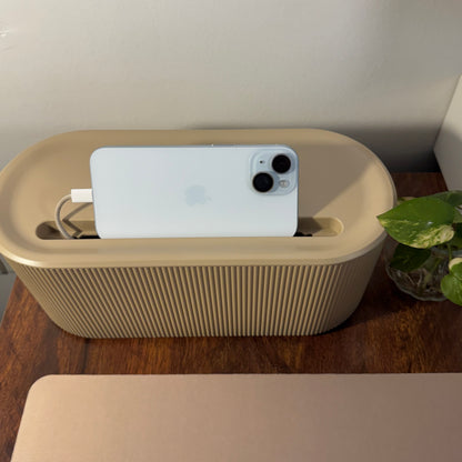 Beige Cable Management box with phone, box, and charger on a wooden surface next to a plant.